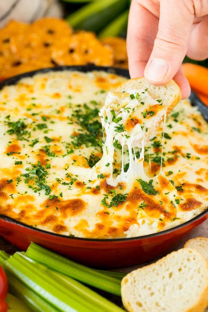 A skillet of artichoke dip with a hand reaching to take a serving.