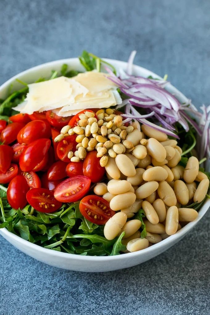 Arugula in a bowl with tomatoes, pine nuts, red onion and shaved parmesan cheese.