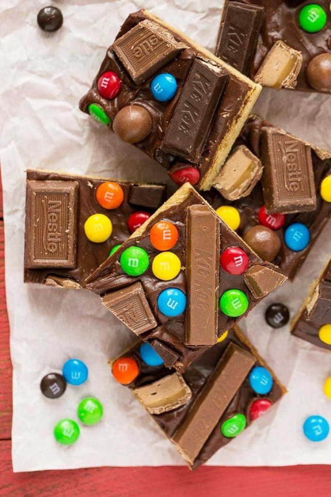 Several candy bar blondies stacked on a paper topped with rich chocolate frosting and a variety of candies.