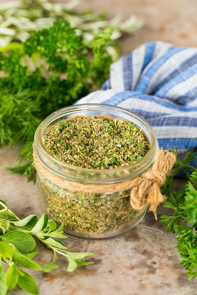 A jar of Greek seasoning surrounded by fresh herbs.