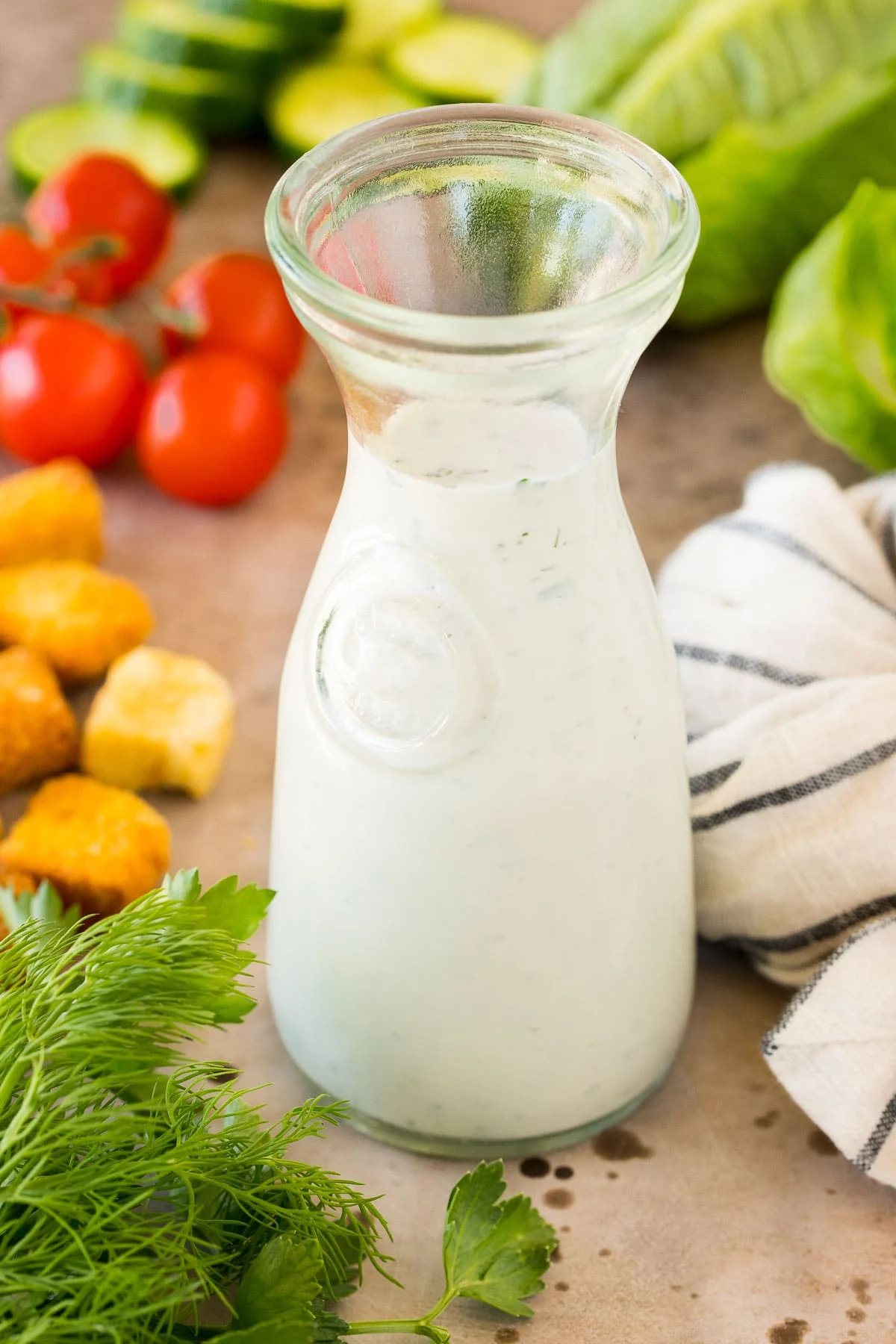 A glass container of kitchen-made ranch dressing surrounded by salad ingredients.