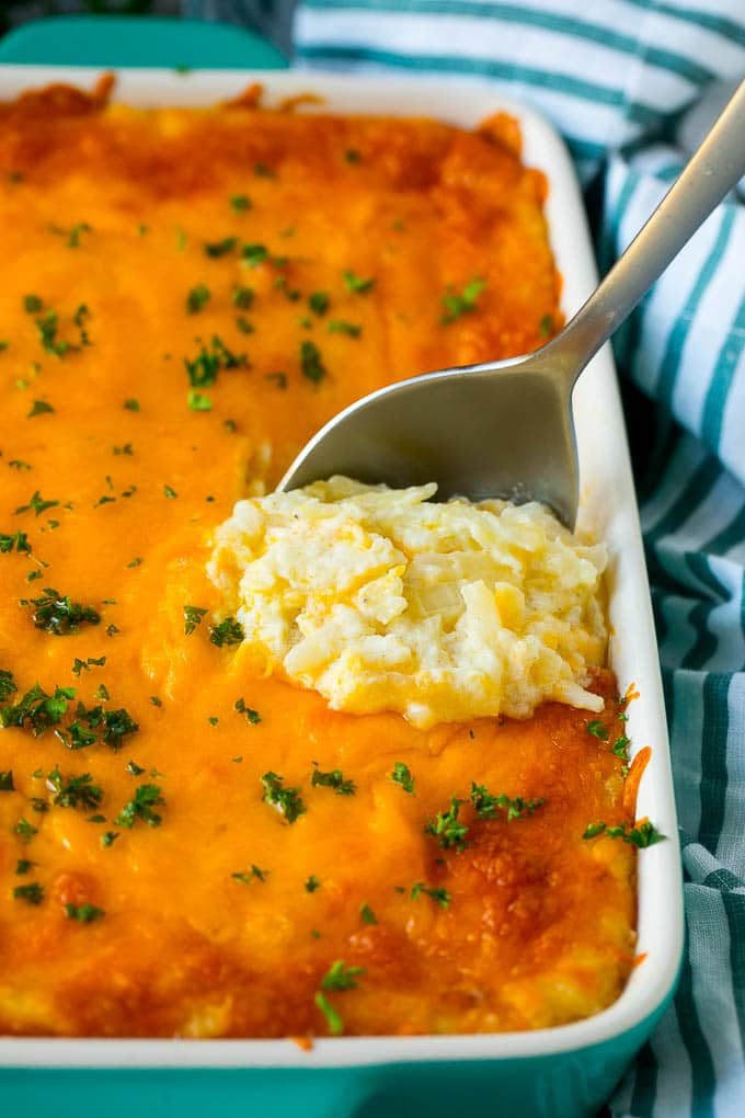 Hash brown casserole with potatoes, cheddar cheese, onions and sour cream, with a serving spoon in the dish.