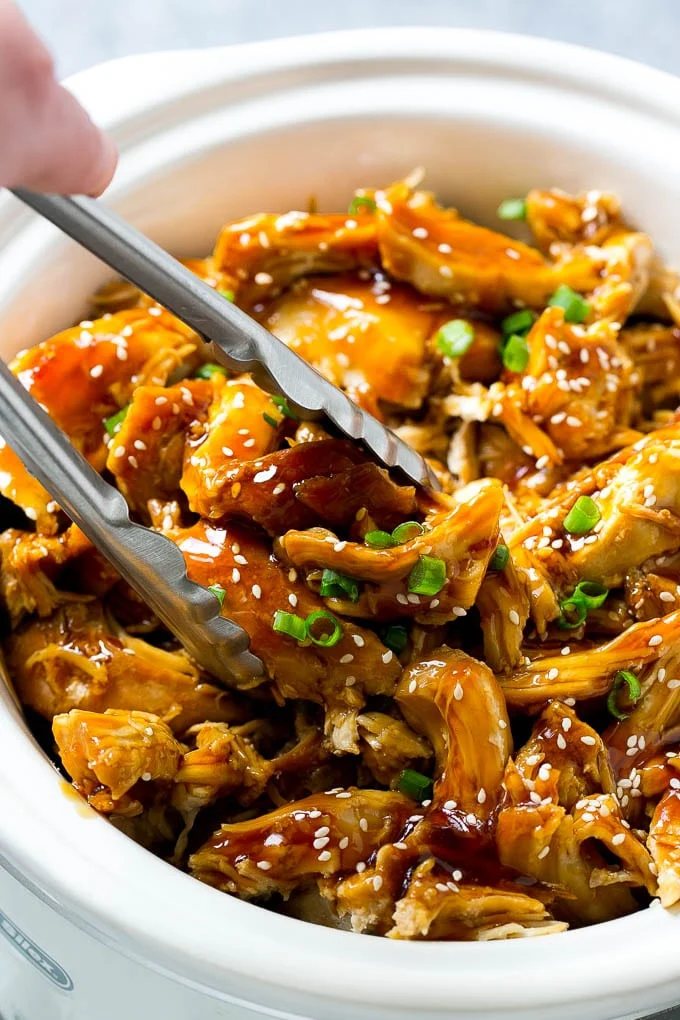 A slow cooker full of teriyaki chicken with tongs in it.