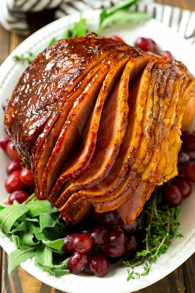 A brown sugar glazed smoked ham on a serving platter surrounded by grapes and fresh herbs.