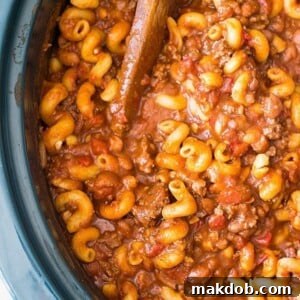 An image of chili mac in a slow cooker with a wood spoon.