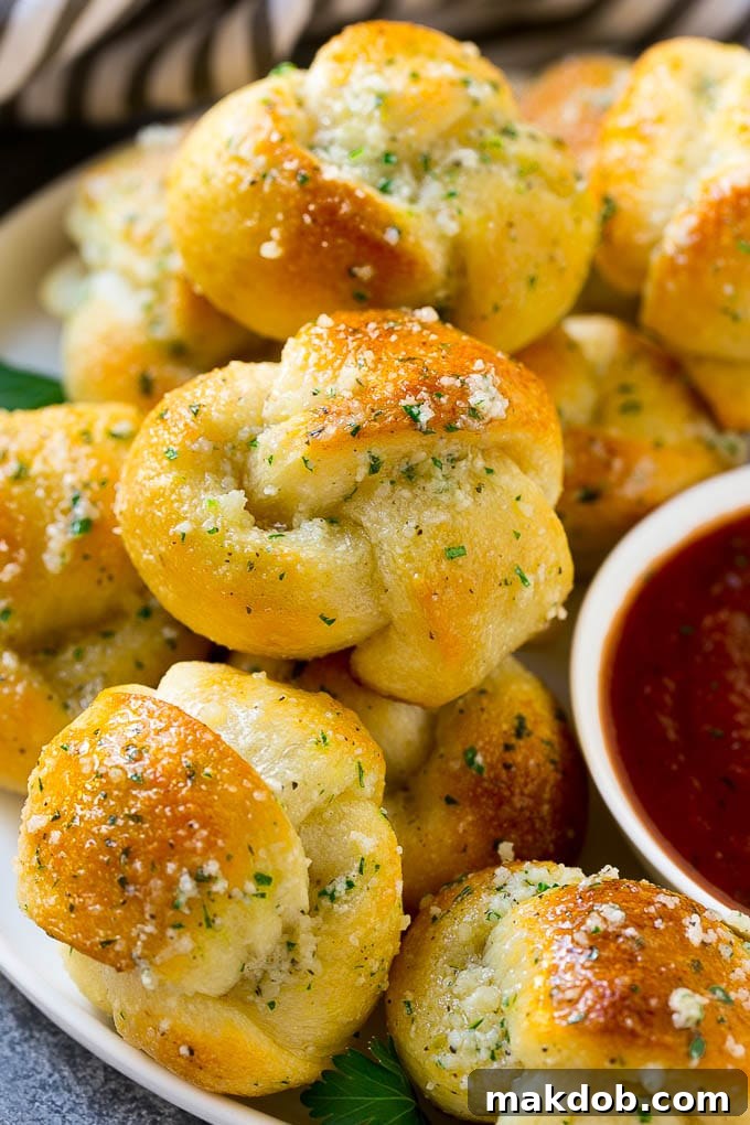 A plate of golden-brown garlic knots slathered in butter and herbs, perfectly baked, next to a small cup of rich marinara sauce for dipping.