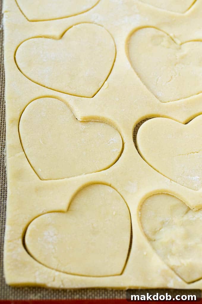 A sheet of perfectly rolled sugar cookie dough with several heart shapes already cut out and a heart cookie cutter nearby.