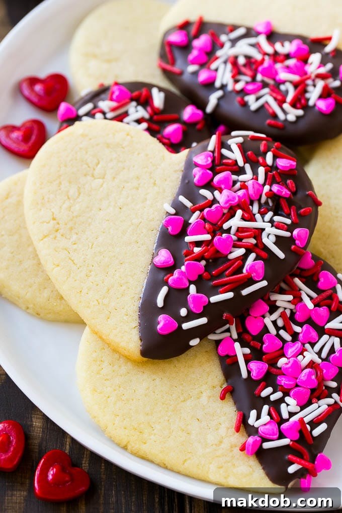 Close-up of heart sugar cookies, half-dipped in dark chocolate and generously coated with colorful Valentine's Day sprinkles, arranged on parchment paper.