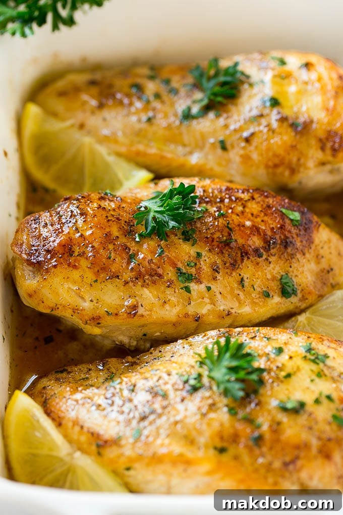 A close-up shot of a perfectly baked lemon chicken breast in a white dish, beautifully garnished with fresh green herbs.