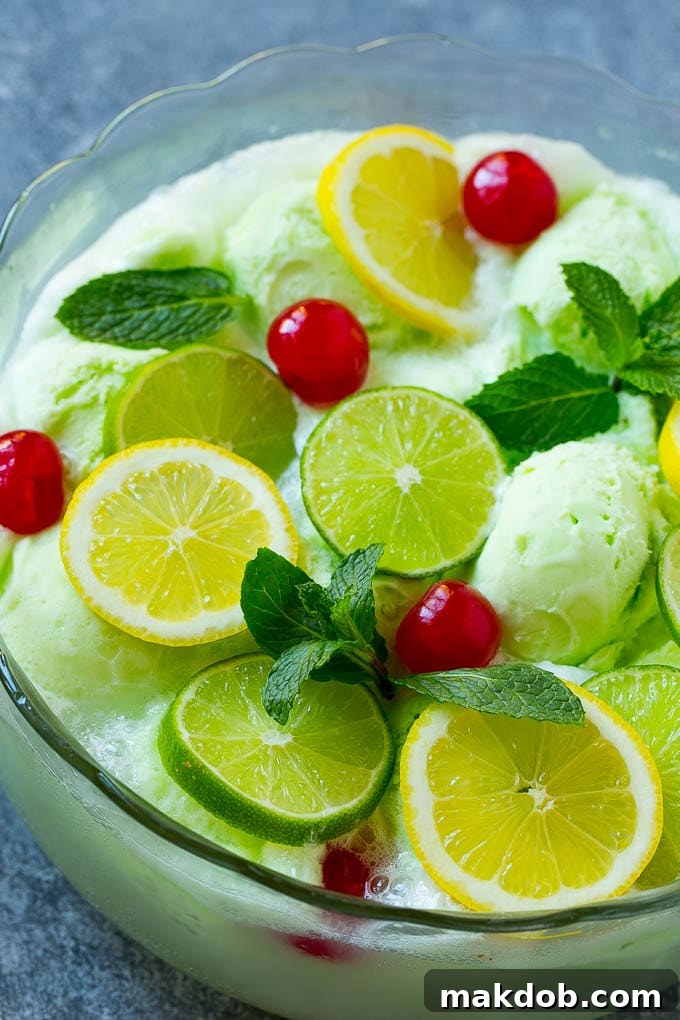 A large punch bowl filled with bright green lime sherbet punch, garnished generously with fresh lime slices, lemon slices, red maraschino cherries, and green mint sprigs, highlighting its refreshing and festive appearance.