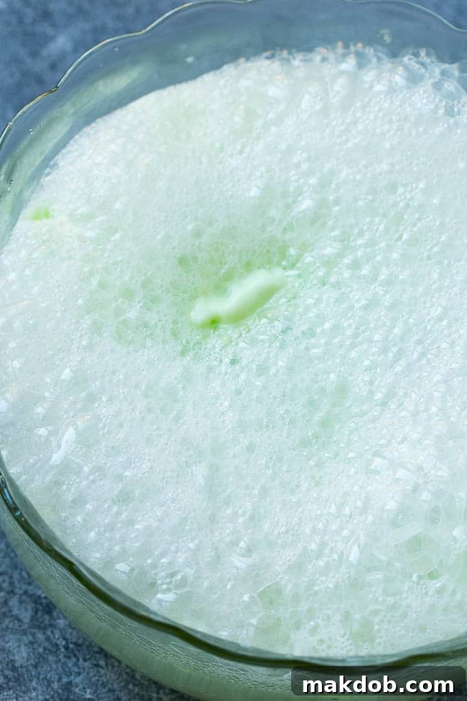 A clear glass punch bowl being filled with sparkling lemon-lime soda, creating an effervescent layer over the creamy limeade and sherbet base.