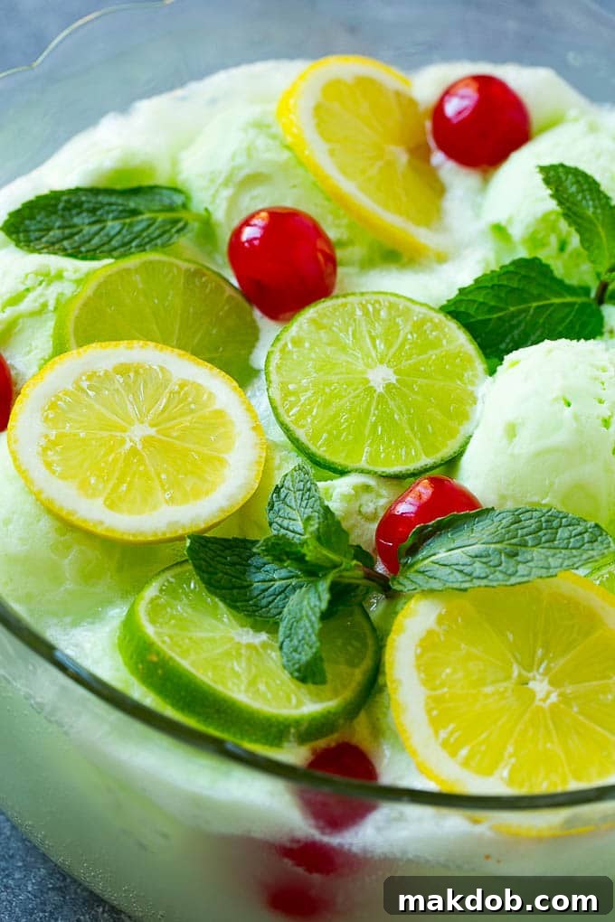 A close-up view of a vibrant lime sherbet punch, showcasing the frothy texture, floating sherbet scoops, and fresh garnishes of lemon, lime, cherries, and mint, inviting a refreshing sip.