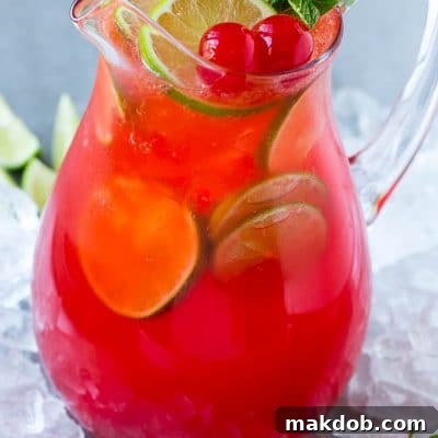 A glass pitcher of cherry limeade garnished with slices of fresh lime and maraschino cherries.