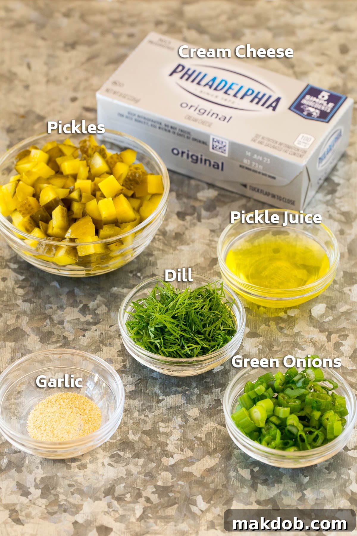 A vibrant flat lay image showcasing the fresh ingredients for dill pickle dip, including a block of cream cheese, a jar of dill pickles, a bunch of fresh dill, green onions, and a small container of garlic powder.