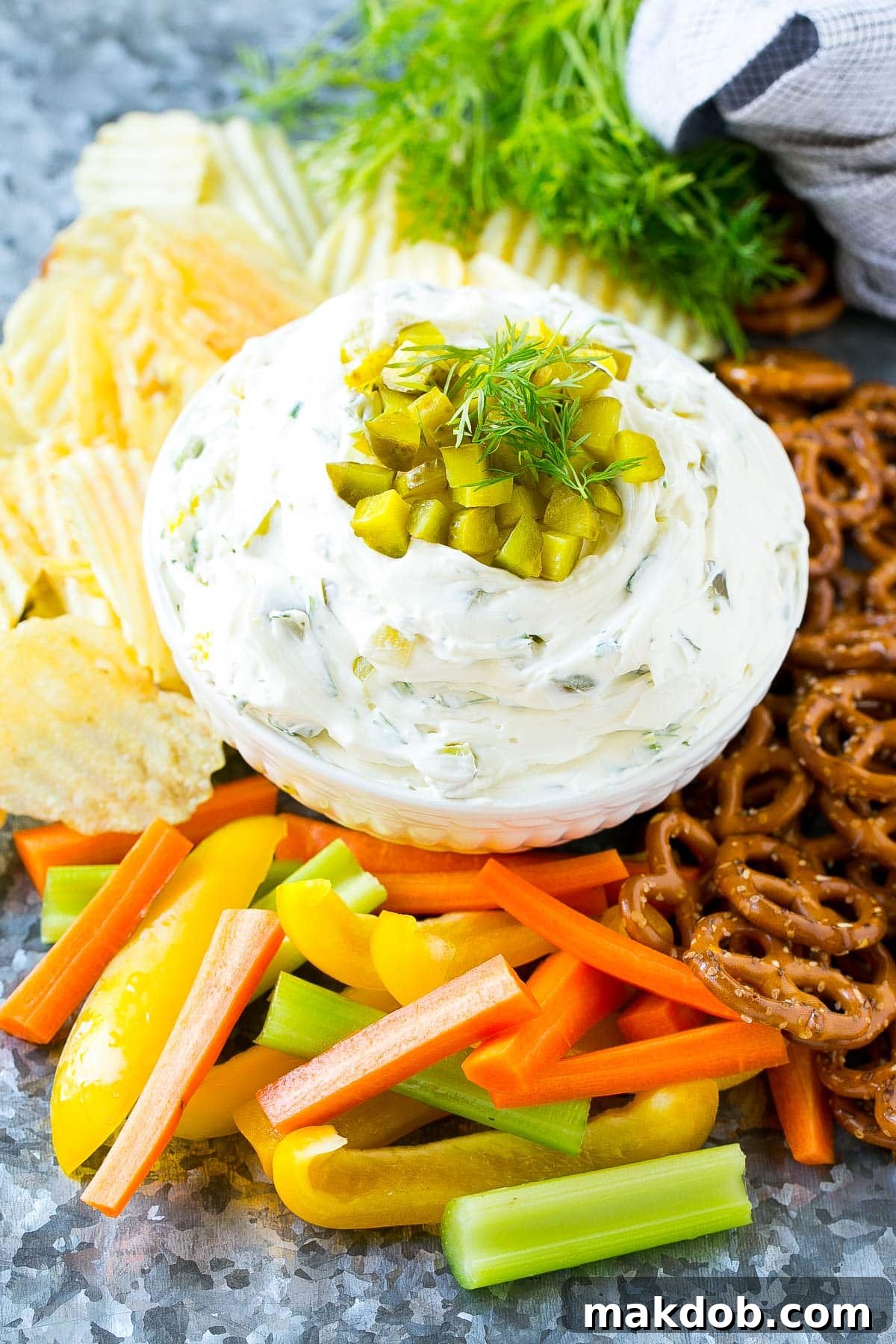 A lavish spread featuring a central bowl of creamy dill pickle dip, surrounded by an abundant display of fresh vegetables like carrots and celery, various crackers, and golden pretzels, creating a perfect party appetizer arrangement.