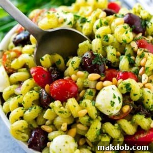 Pesto pasta salad featuring sweet cherry tomatoes, briny olives, creamy mozzarella balls, crunchy pine nuts, and thinly sliced red onion.