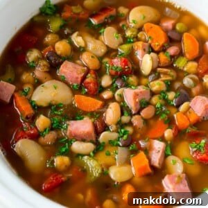 A slow cooker brimming with hearty ham and bean soup, made with a flavorful blend of vegetables, dried beans, and diced ham.