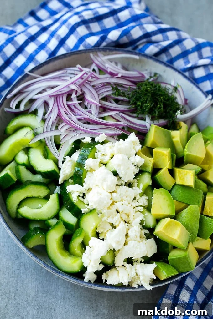 A vibrant collection of fresh salad ingredients, including sliced cucumbers, red onion, avocado, fresh dill, and crumbled feta cheese, beautifully arranged in a large mixing bowl.