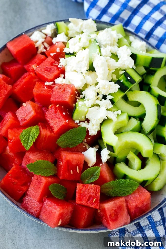 Close-up of a bowl containing perfectly cubed watermelon, thinly sliced cucumbers, crumbled white feta cheese, and vibrant green mint leaves, ready to be dressed and served.