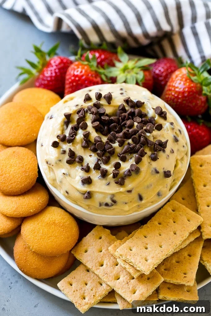 A pristine bowl of irresistible cookie dough dip, generously crowned with an abundance of mini chocolate chips and surrounded by an inviting assortment of dippers, including various cookies and fresh, vibrant fruit.
