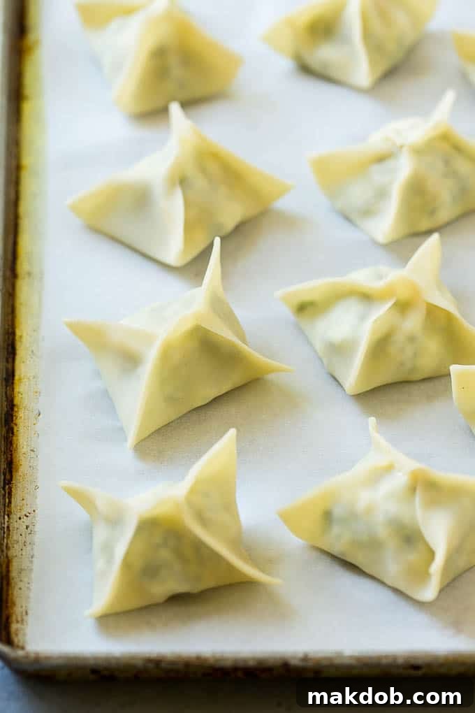 Neatly folded spinach artichoke wontons arranged on a baking sheet, ready for frying.