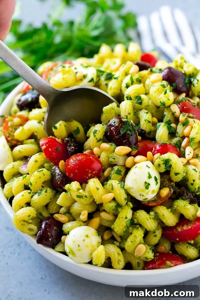 A vibrant pesto pasta salad featuring corkscrew pasta, halved cherry tomatoes, fresh mozzarella balls, Kalamata olives, toasted pine nuts, and finely diced red onion, all generously coated in green basil pesto.