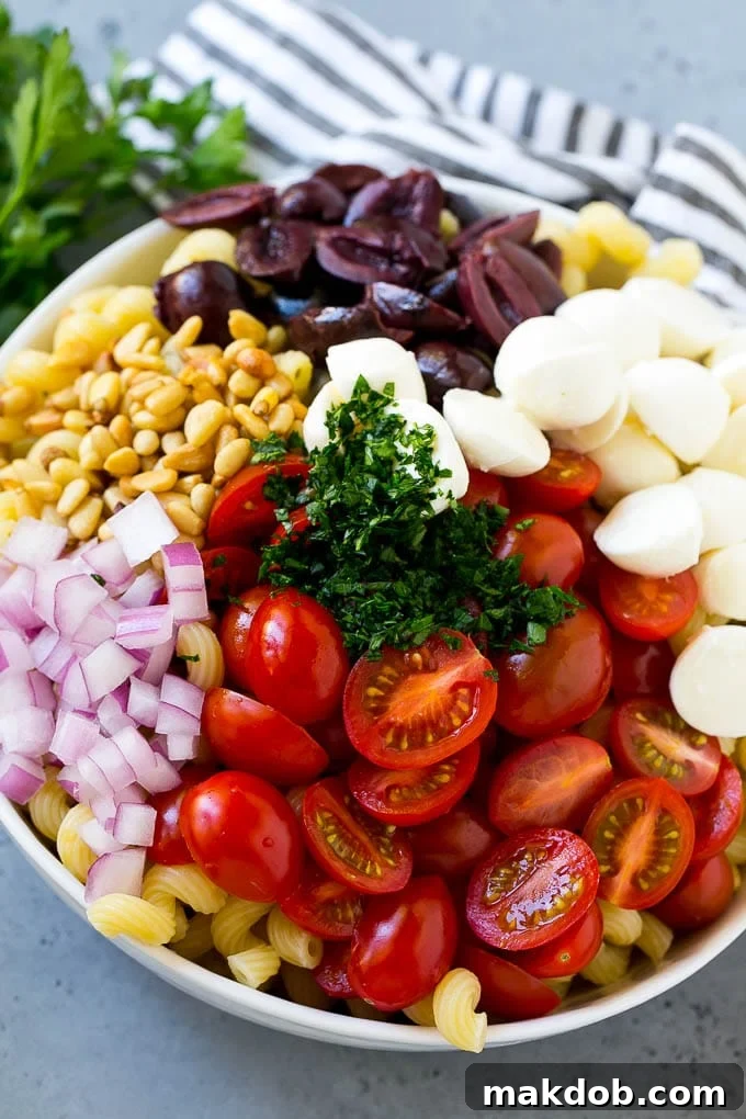 A close-up shot of the pesto pasta salad ingredients before mixing, featuring corkscrew pasta, bright red cherry tomatoes, creamy white mozzarella balls, dark Kalamata olives, green parsley, and thinly sliced red onion.