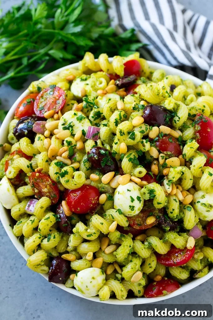 A close-up of a bowl of freshly prepared pesto pasta salad, beautifully garnished with a sprinkle of toasted pine nuts and finely chopped fresh parsley on top.