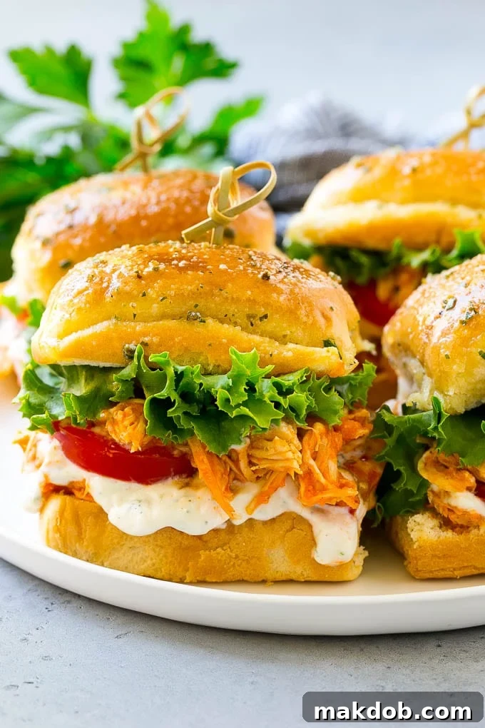 A plate of freshly prepared Buffalo Chicken Sliders, garnished with vibrant green lettuce, bright red tomato slices, and a creamy drizzle of ranch dressing.