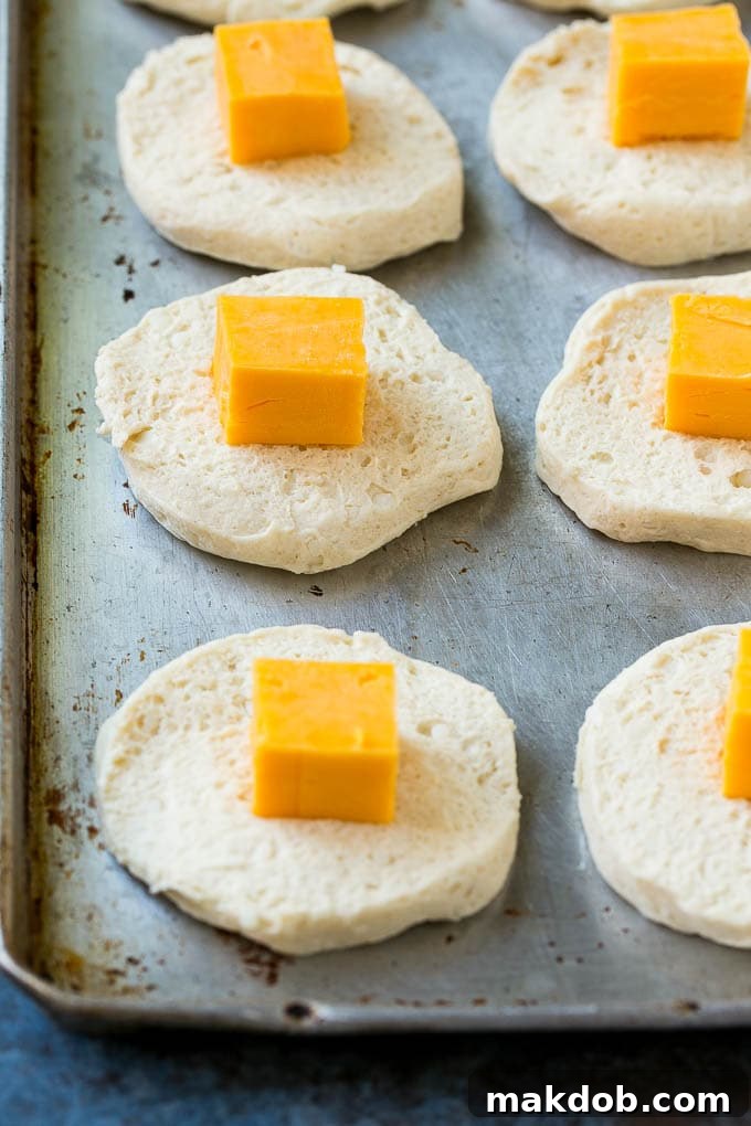 Rounds of uncooked biscuit dough, each topped with a single cube of sharp cheddar cheese, laid out on a baking sheet, ready for assembly.