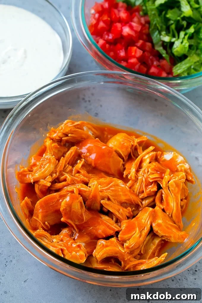 An overhead view showing a bowl of buffalo-sauced shredded chicken, surrounded by small bowls of ranch dressing, diced tomatoes, and shredded lettuce, ready for taco assembly.