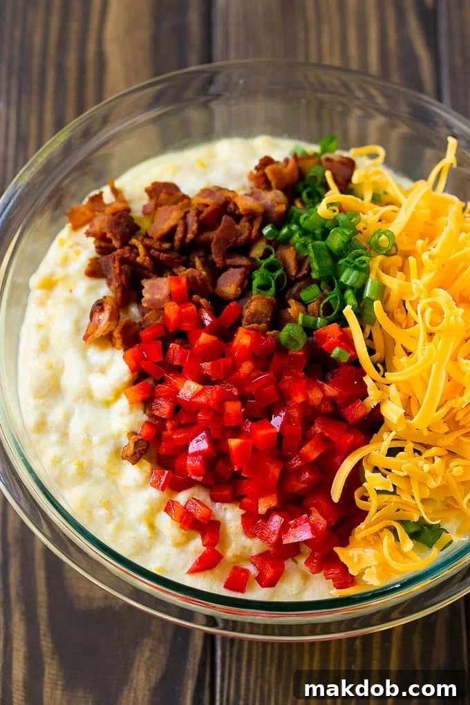 Loaded Corn Casserole Recipe | Easy Corn Casserole | Bacon Corn Casserole | Corn Pudding #corn #casserole #bacon #cheese #sidedish #thanksgiving #dinner #dinneratthezoo A vibrant bowl of corn casserole batter, studded with pieces of red bell pepper, green onions, shredded cheddar cheese, and crispy bacon.