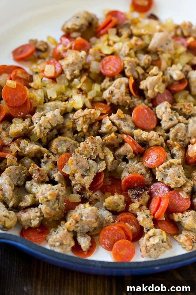 Close-up of browned Italian sausage crumbles and finely diced onions sizzling in a large skillet, forming the flavorful base for the pizza pasta.
