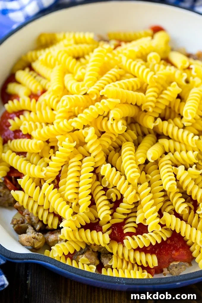 Uncooked rotini pasta nestled in a skillet with browned sausage, pepperoni, and rich tomato sauce, ready to be simmered into a delicious one-pot meal.