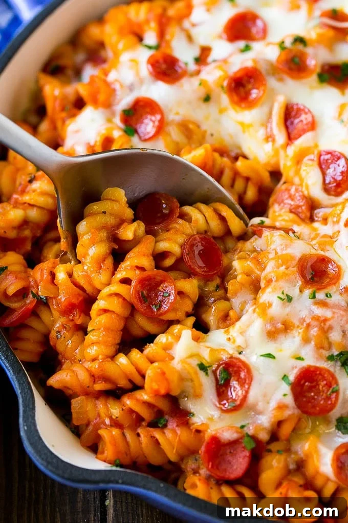 A large skillet brimming with hot, cheesy pizza pasta, featuring tender rotini, savory sausage, and pepperoni, topped with melted mozzarella. A serving spoon is resting in the pan, ready for dishing out.