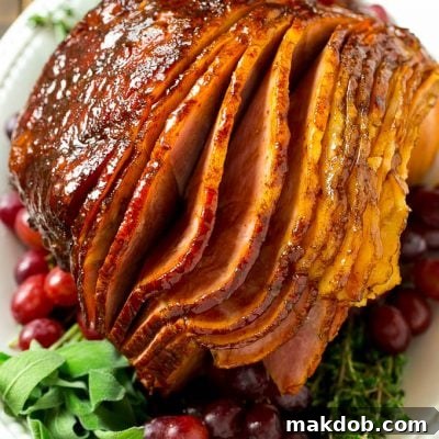 A brown sugar glazed smoked ham on a serving platter surrounded by grapes and fresh herbs.