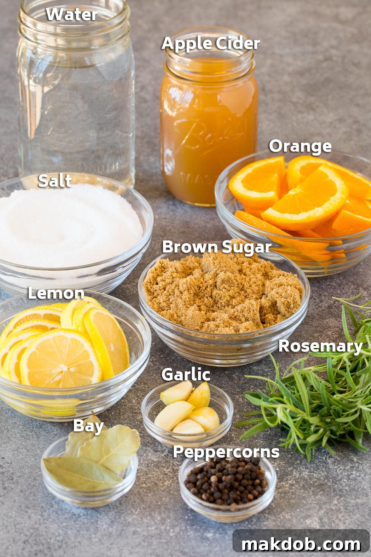 Various turkey brine ingredients displayed, including a jug of apple cider, bowls of salt and brown sugar, and fresh herbs like rosemary and citrus slices.