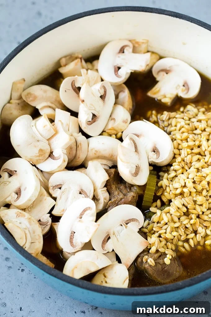 A pot of beef barley soup with sliced mushrooms and barley stirred in, continuing to simmer.