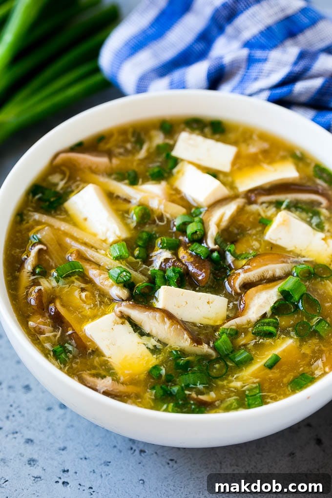 A steaming bowl of Hot and Sour Soup, artfully garnished with green onions, showcasing a rich broth filled with tofu cubes, mushroom slices, and delicate egg ribbons, ready to be enjoyed as a classic Asian comfort food.