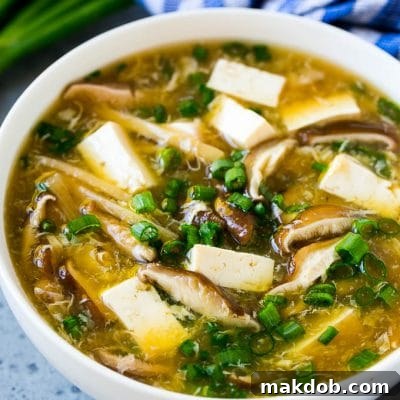 A bowl of hot and sour soup filled with tofu, mushrooms, egg, bamboo shoots and green onion.