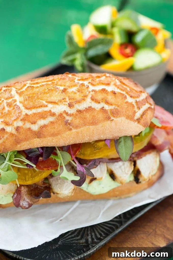 A close-up of a generously filled sandwich roll with slices of grilled chicken, crisp bacon, fresh tomatoes, and leafy greens.