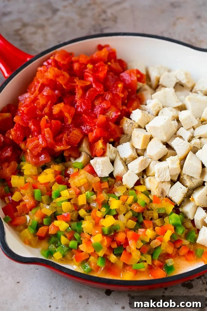 Cubed chicken, cooked bell peppers, and tomatoes simmering in a skillet, forming the creamy filling for the casserole.