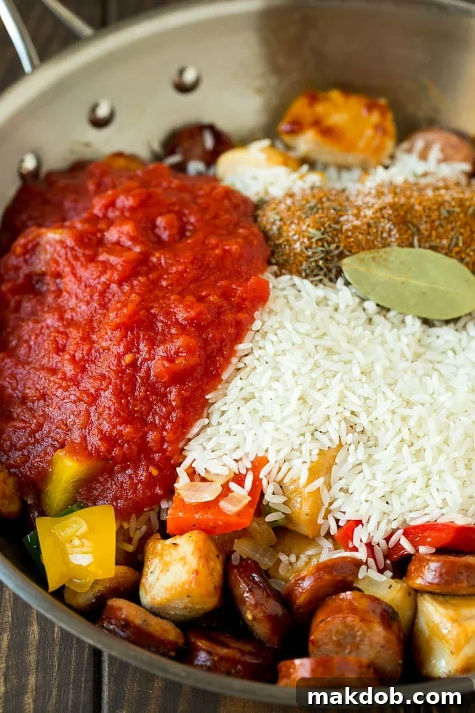 A mix of vegetables, chicken, sausage, rice, and crushed tomatoes simmering together in a large pan, forming a delicious jambalaya base.