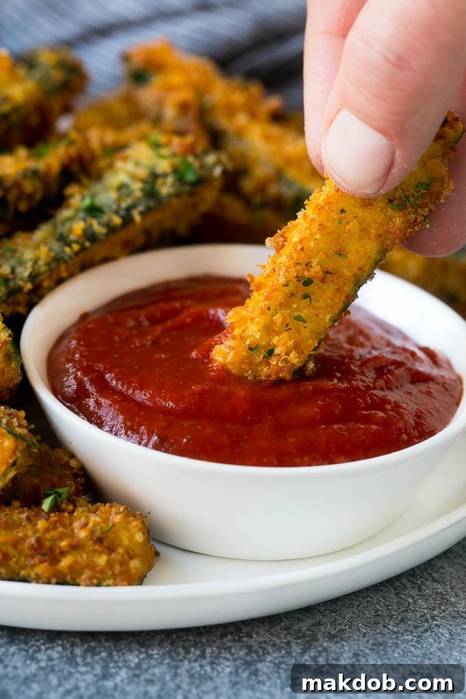A hand dipping a piece of crispy fried zucchini into a bowl of vibrant marinara sauce.
