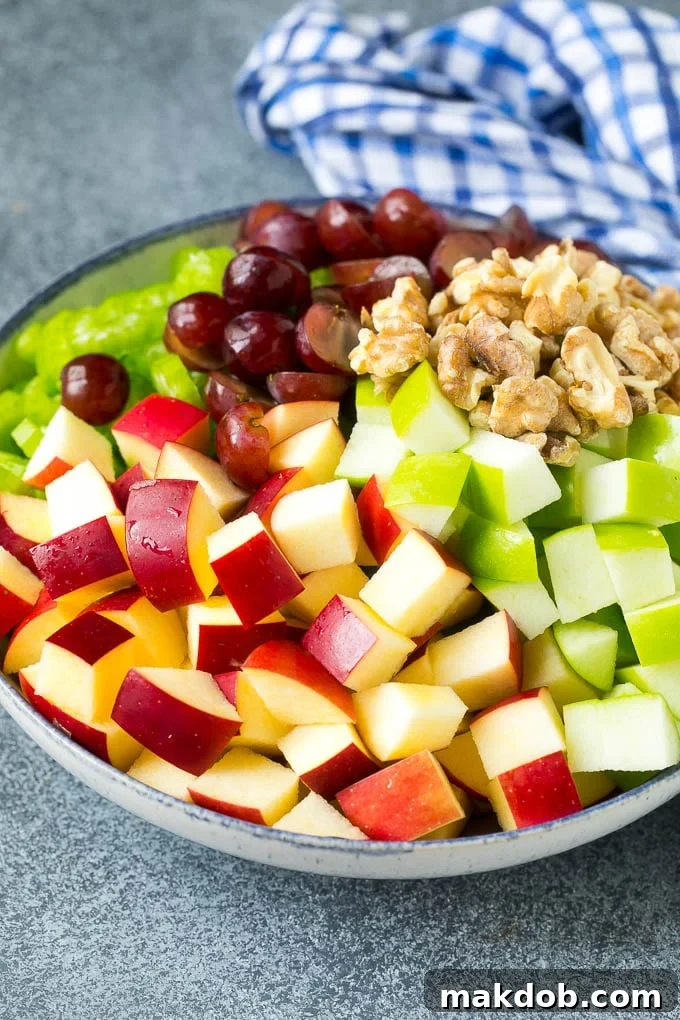 A vibrant mix of red and green apples, dark red grapes, crisp green celery stalks, and golden walnuts, all diced and ready to be tossed in a creamy Waldorf salad dressing, presented in a large glass serving bowl.