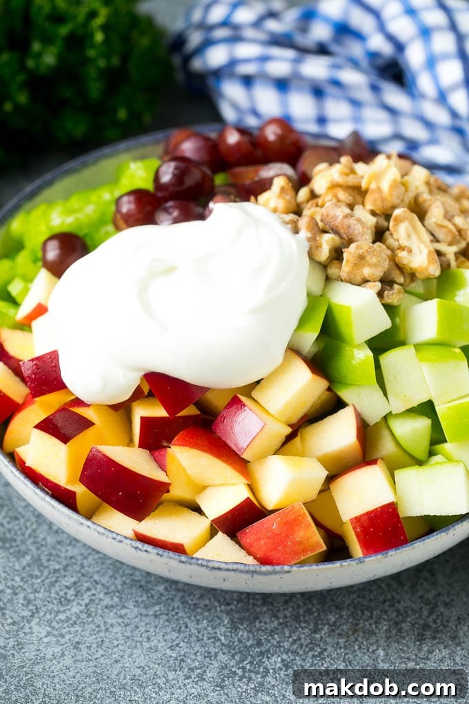 Crisp red and green apples, sweet grapes, crunchy celery, and rich walnuts, all diced and combined in a bowl, awaiting the pour of a creamy, homemade Waldorf dressing, highlighting the fresh ingredients.