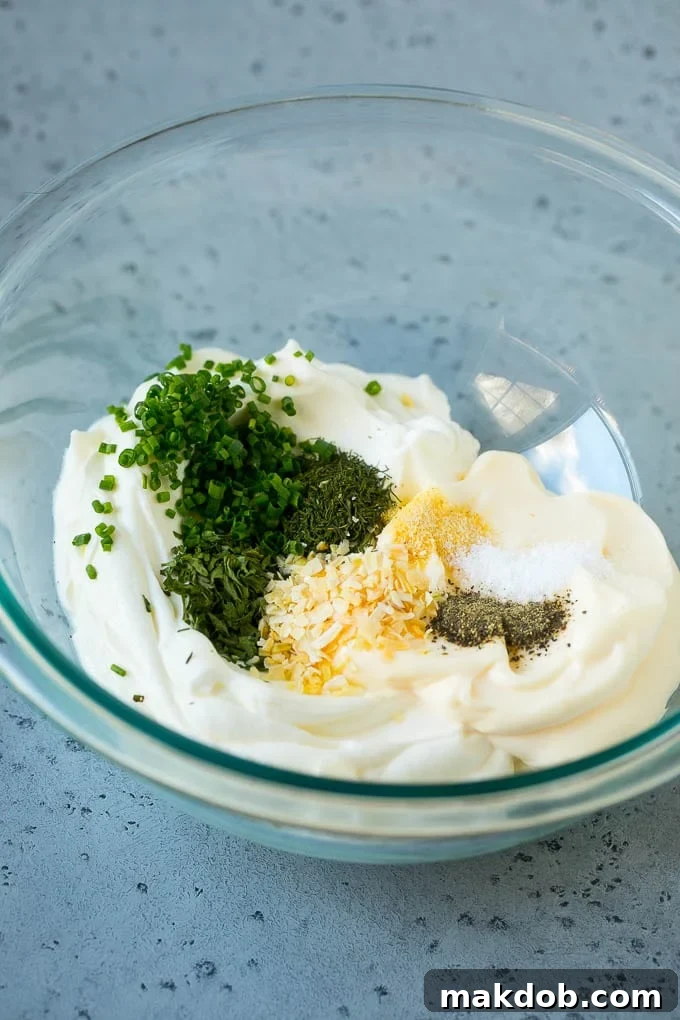 Garden Herb Dip 3 A close-up shot of a mixing bowl containing the primary ingredients for veggie dip: creamy sour cream and mayonnaise, speckled with a variety of dried and fresh herbs and spices, ready to be combined.