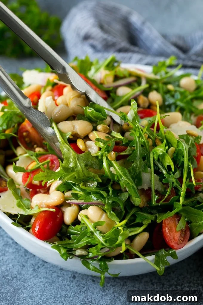 Arugula Salad Recipe | Green Salad | White Bean Salad #salad #arugula #cleaneating #lowcarb #keto #dinneratthezoo Tongs gracefully serving a portion of fresh arugula salad from a bowl, highlighting its inviting components.