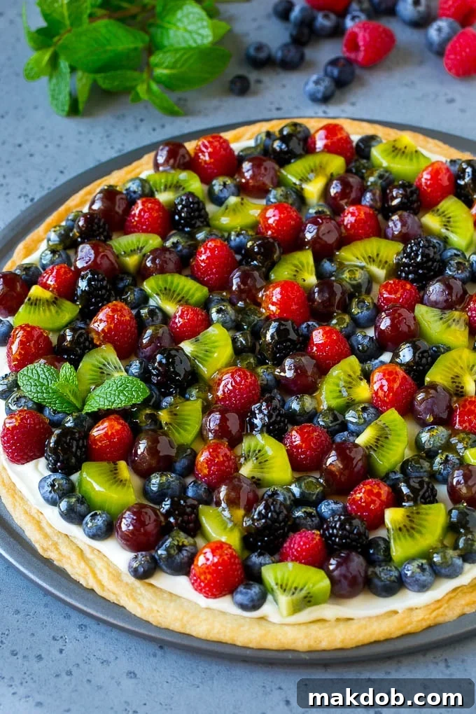 Classic Fruit Pizza Recipe: Sugar Cookie Crust, Cream Cheese Frosting & Fresh Fruit Fruit pizza with a sugar cookie crust and cream cheese frosting, topped with berries, kiwi and grapes, on a white serving platter.