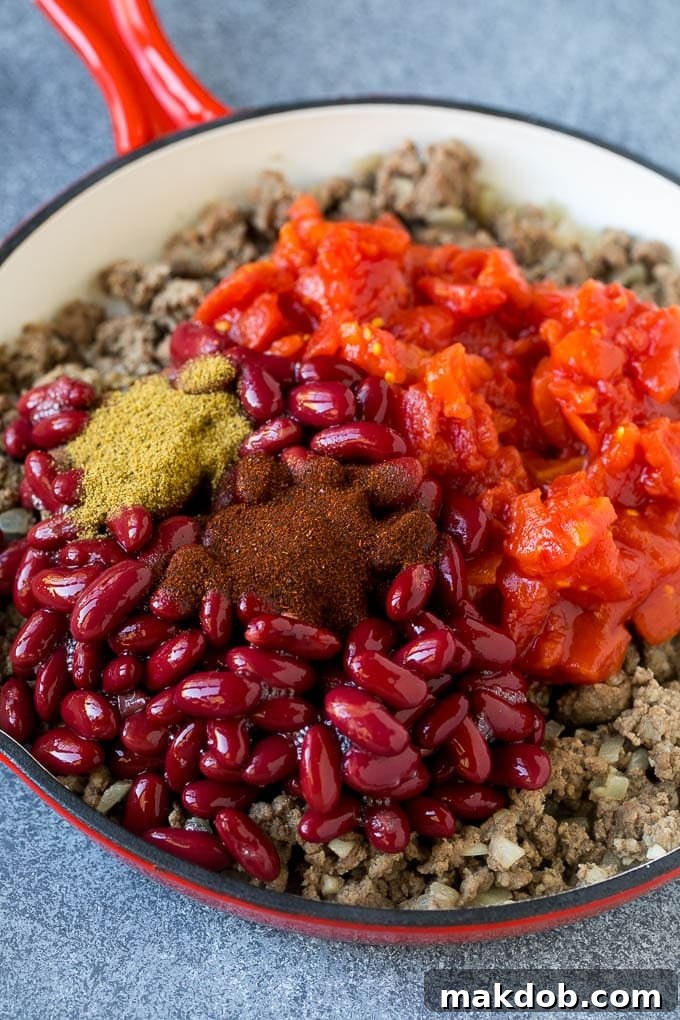 Ground beef, kidney beans, tomatoes and spices simmering in a skillet, capturing the start of the one-pot chili mac.